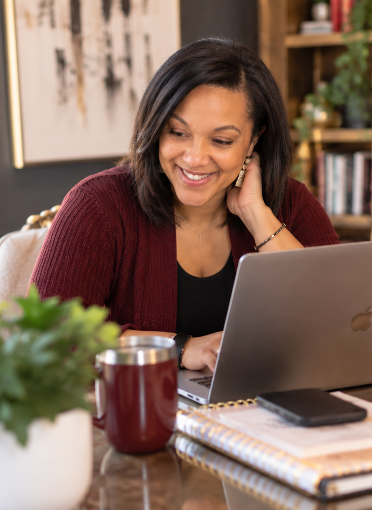 Kristen Moore at her desk, smiling as she works on a laptop with a maroon tumbler and notebook beside her